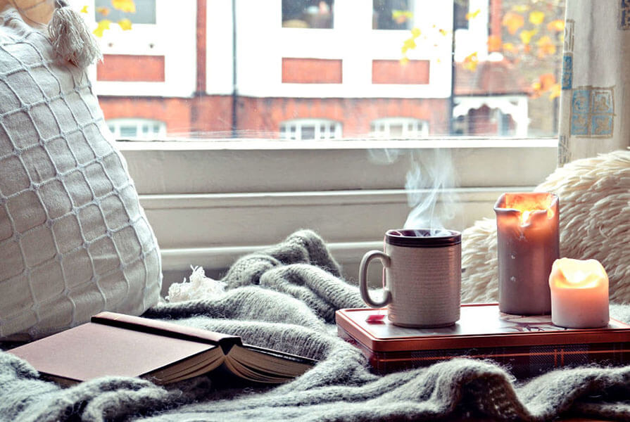 xícara de café ao lado de velas com livro aberto em cima da cama com vista para a rua