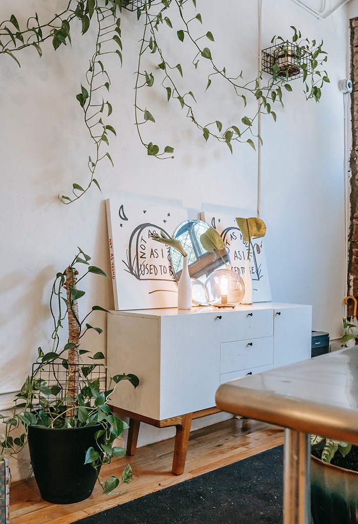 Cômoda estilo aparador com dois quadros apoiados e um espelho redondo ao meio, dois vasos de plantas com folhas largas decoração afetiva