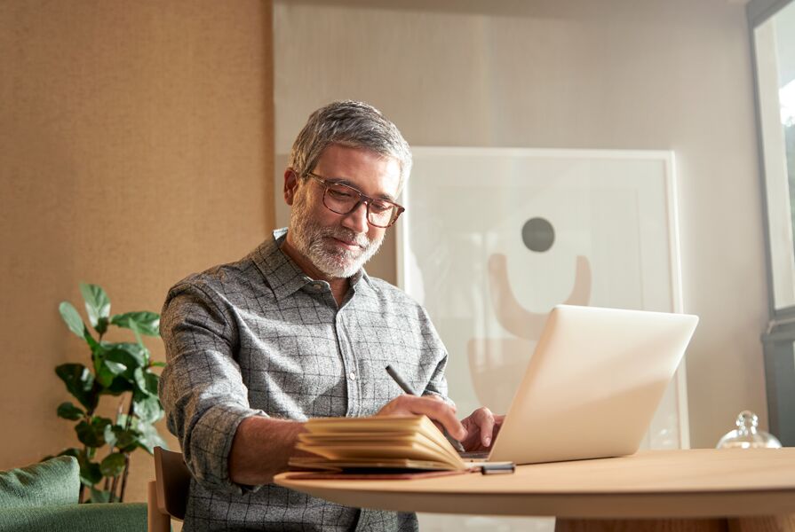 Homem escrevendo em caderno enquanto usa laptop
